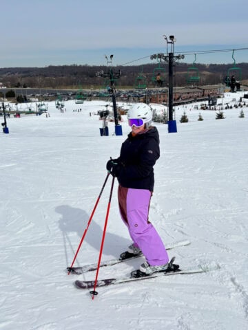 Woman wearing snow pants, jacket and goggles while posing on skis at top of snowy slope.