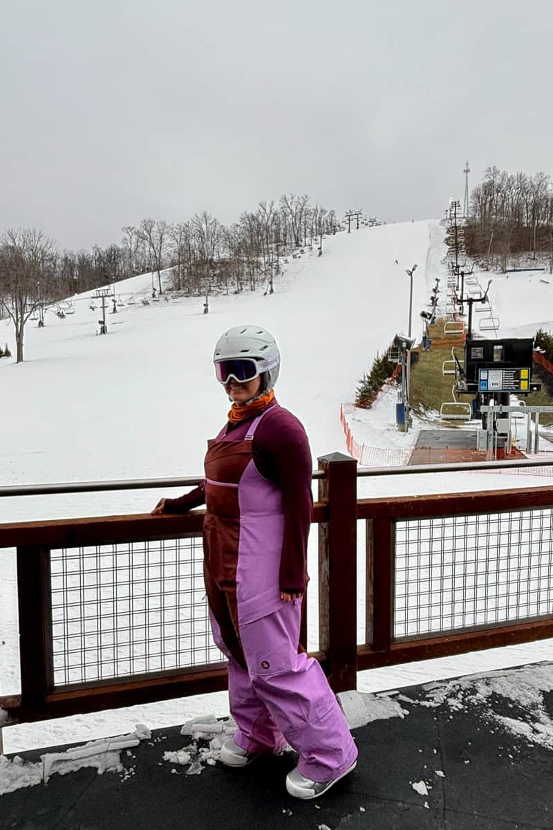Woman wearing purple snow jumper and posing on ski resort deck above snow slopes.
