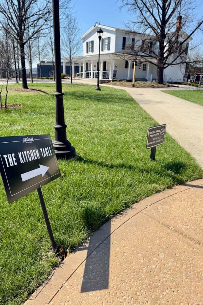 Sign pointing out the direction to take towards The Kitchen Table restaurant on sidewalk passing white two-story house at Jim Beam Distillery.