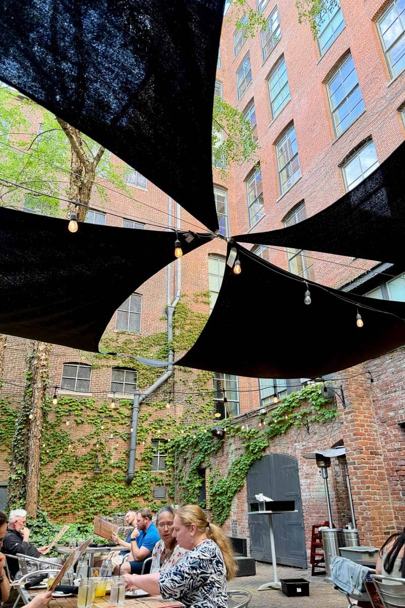 Outdoor dining area in courtyard between brick buildings with canvas shelter and lights stretched out.
