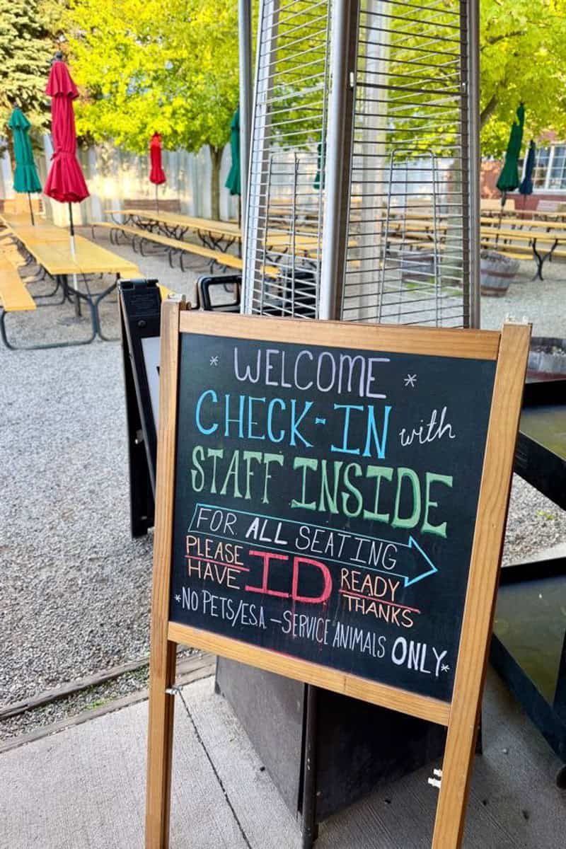 Chalkboard sign near outdoor dining area directing visitors to check in with staff inside.