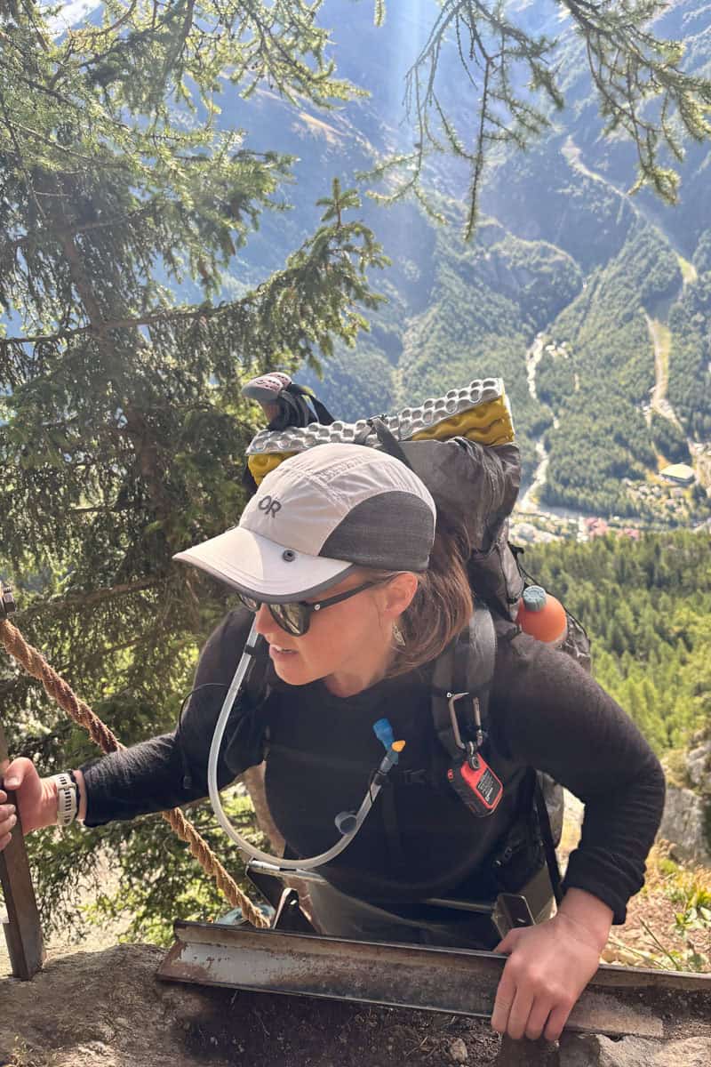 Woman in black wool sun hoodie and ball cap climbing ladder attached to side of hill.