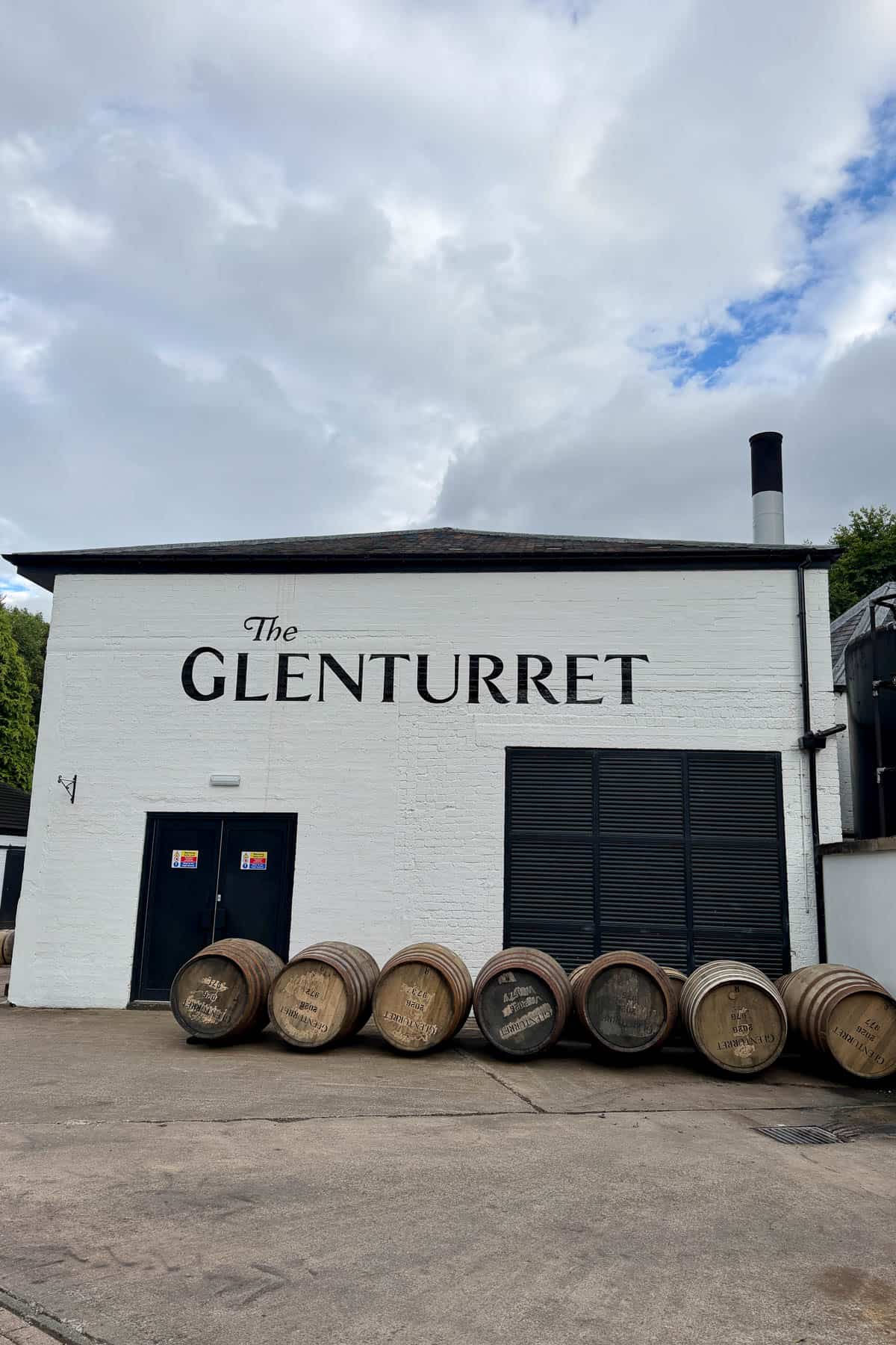 Exterior of Glenturret Distillery, a whitewashed building with black trim and a row of barrels outside.