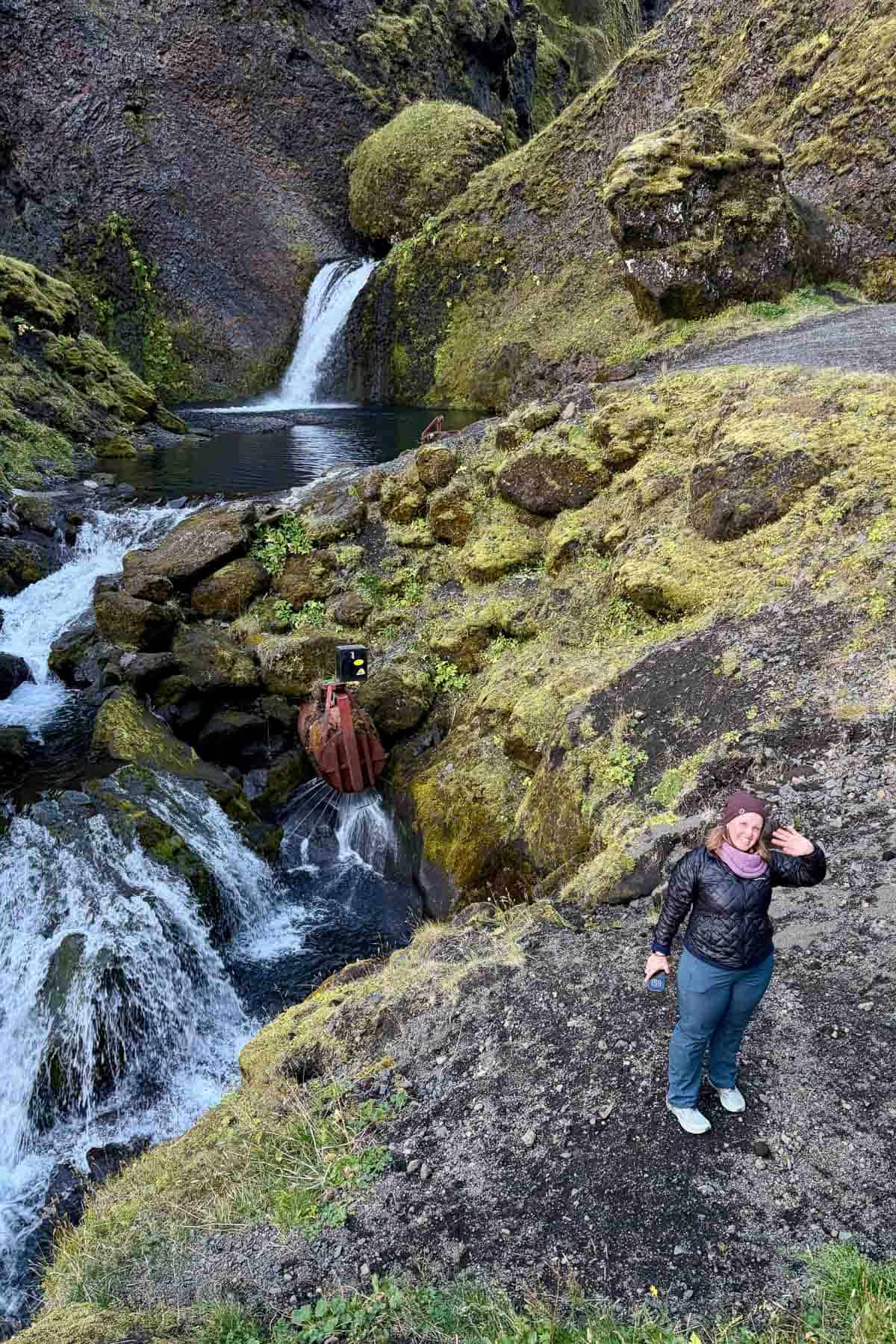 Woman wearing ultralight down jacket and waving while poised on a black sandbank with a waterfall in the background.